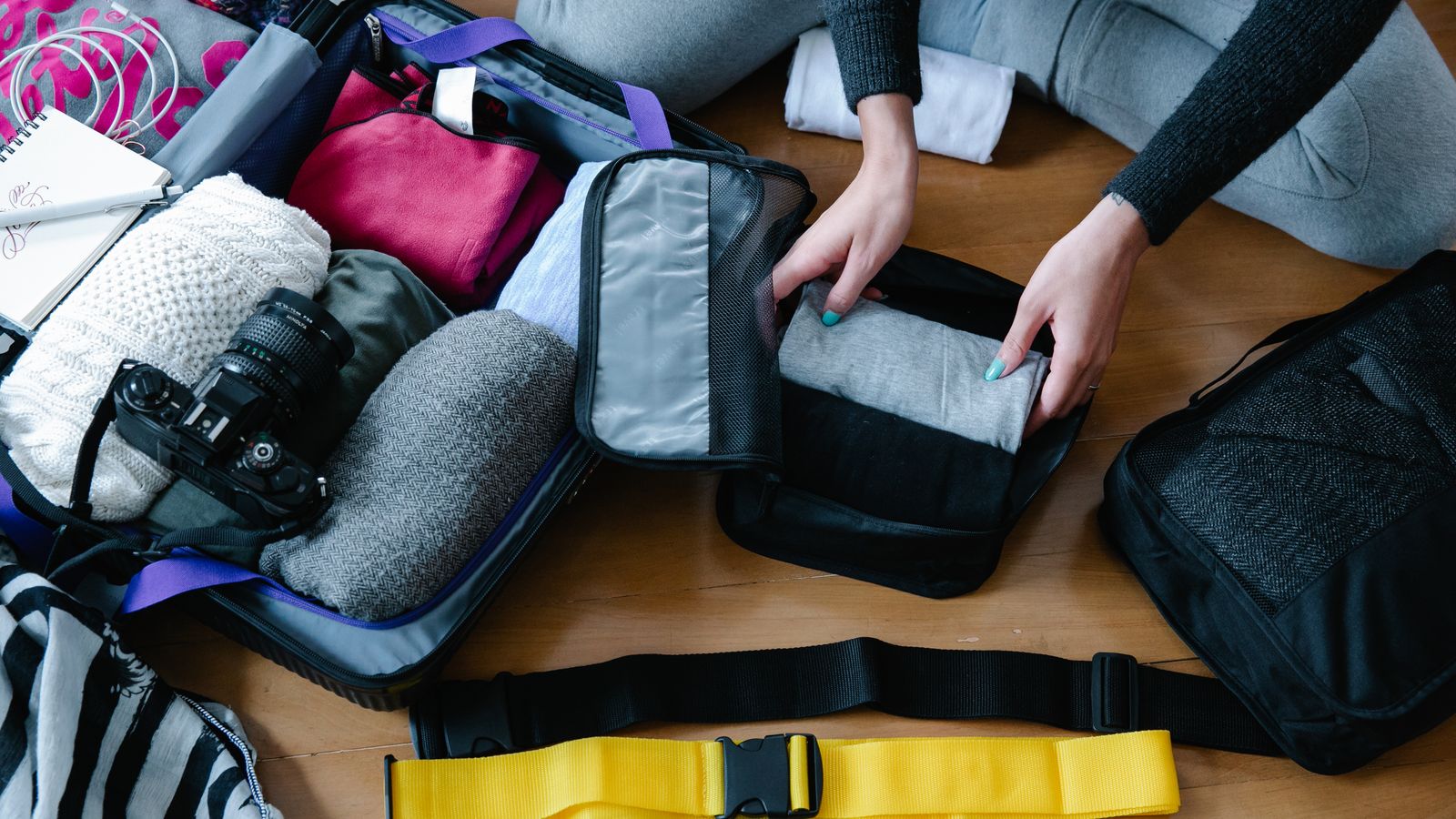 Hands arranging clothes and a camera inside an open cabin bag on a wooden floor